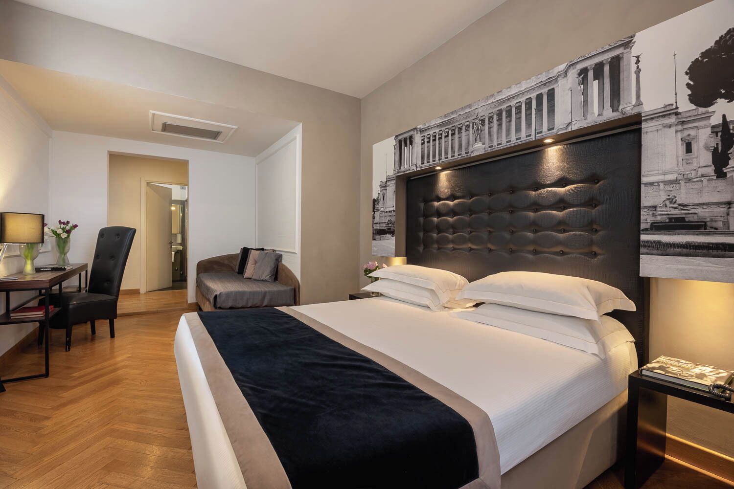 A hotel room with a large bed, a desk with a chair, and a nightstand with a lamp.