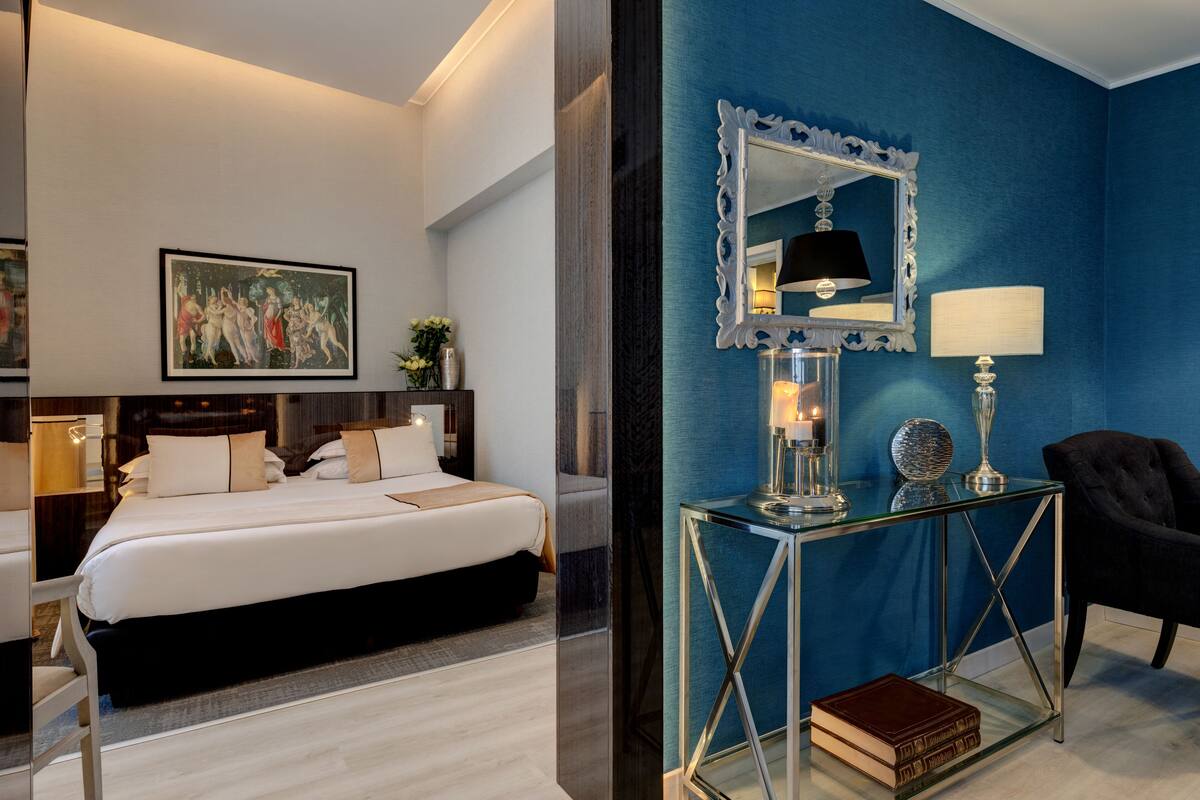 A modern hotel room with a large bed, a glass console table with decorative items, a mirror, and a blue accent wall.