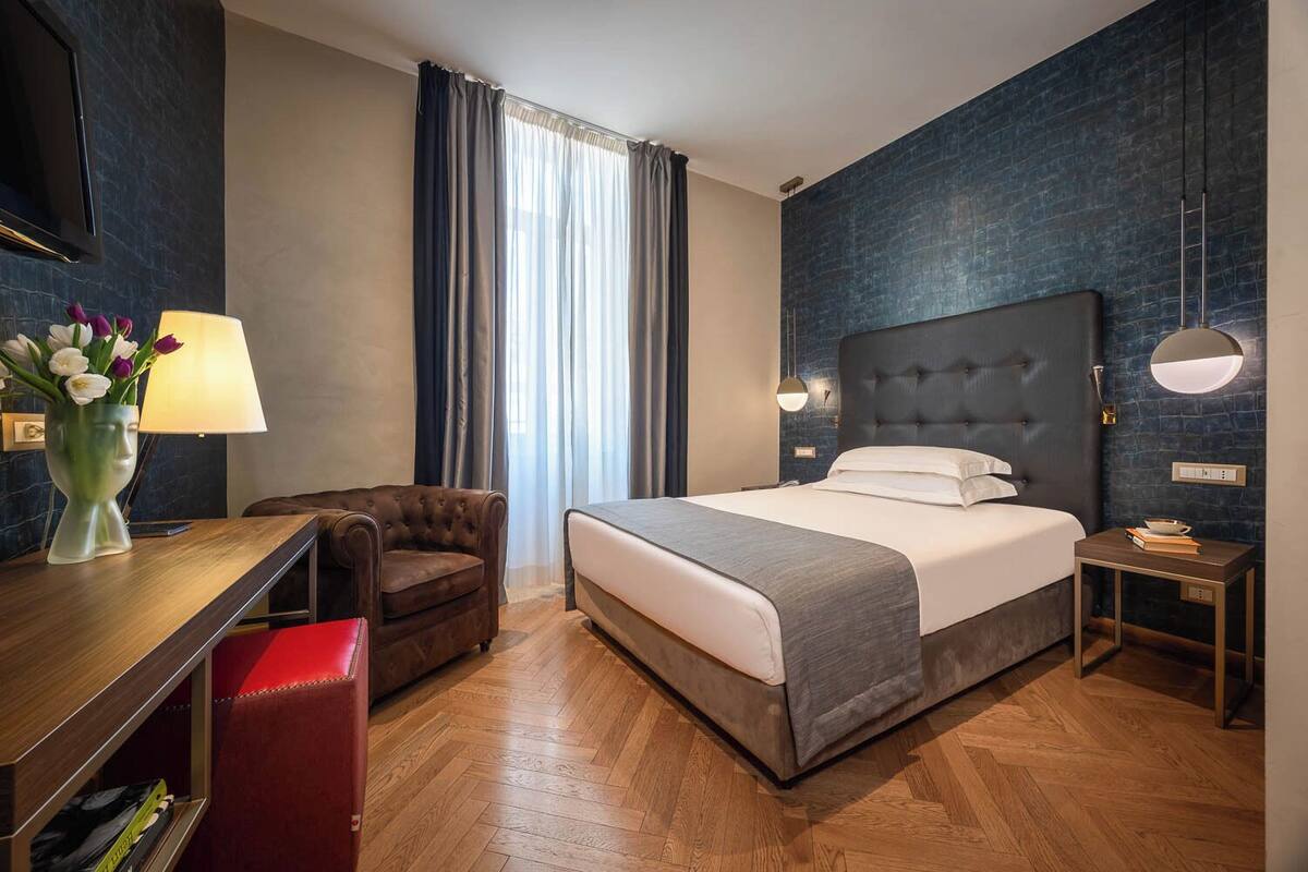 A hotel room with a bed, a desk, a chair, a nightstand, and a wall-mounted TV.