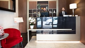Un espace d’accueil d’hôtel au design moderne, comprenant un comptoir d’accueil, un fauteuil rouge, un lustre et un grand miroir mural.