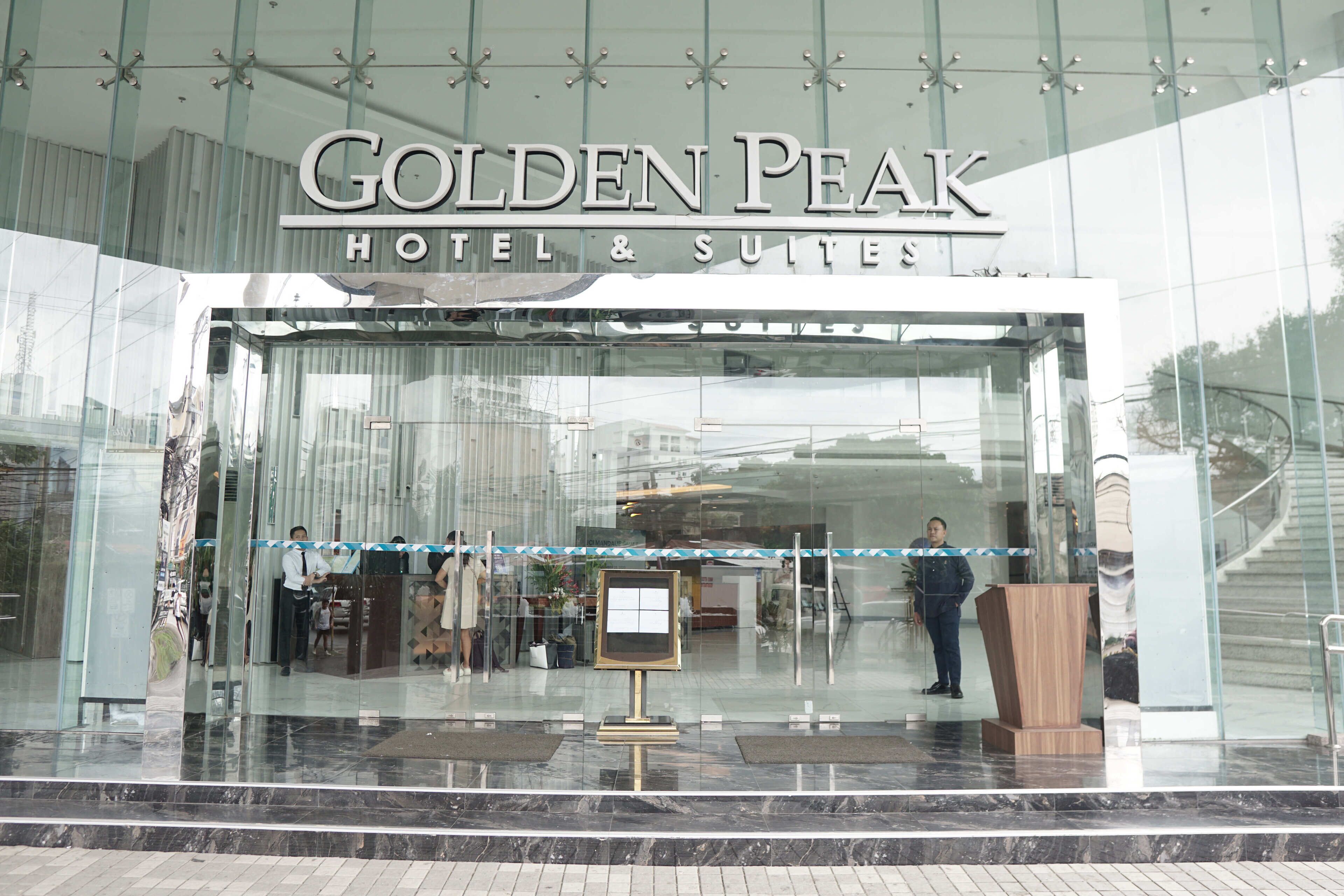 The exterior of the Golden Peak Hotel & Suites with a glass facade, a reception desk, and a signboard.