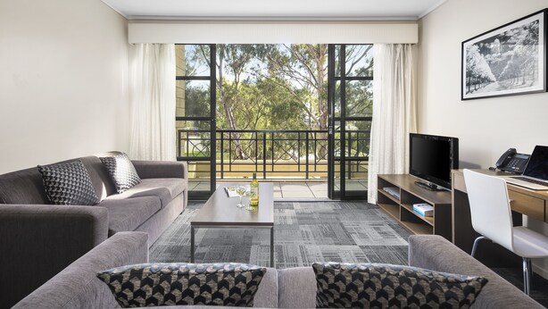 A modern living room with a grey sofa, a coffee table, a TV, and a balcony with a view of trees. - Quest Narre Warren (Narre Warren)
