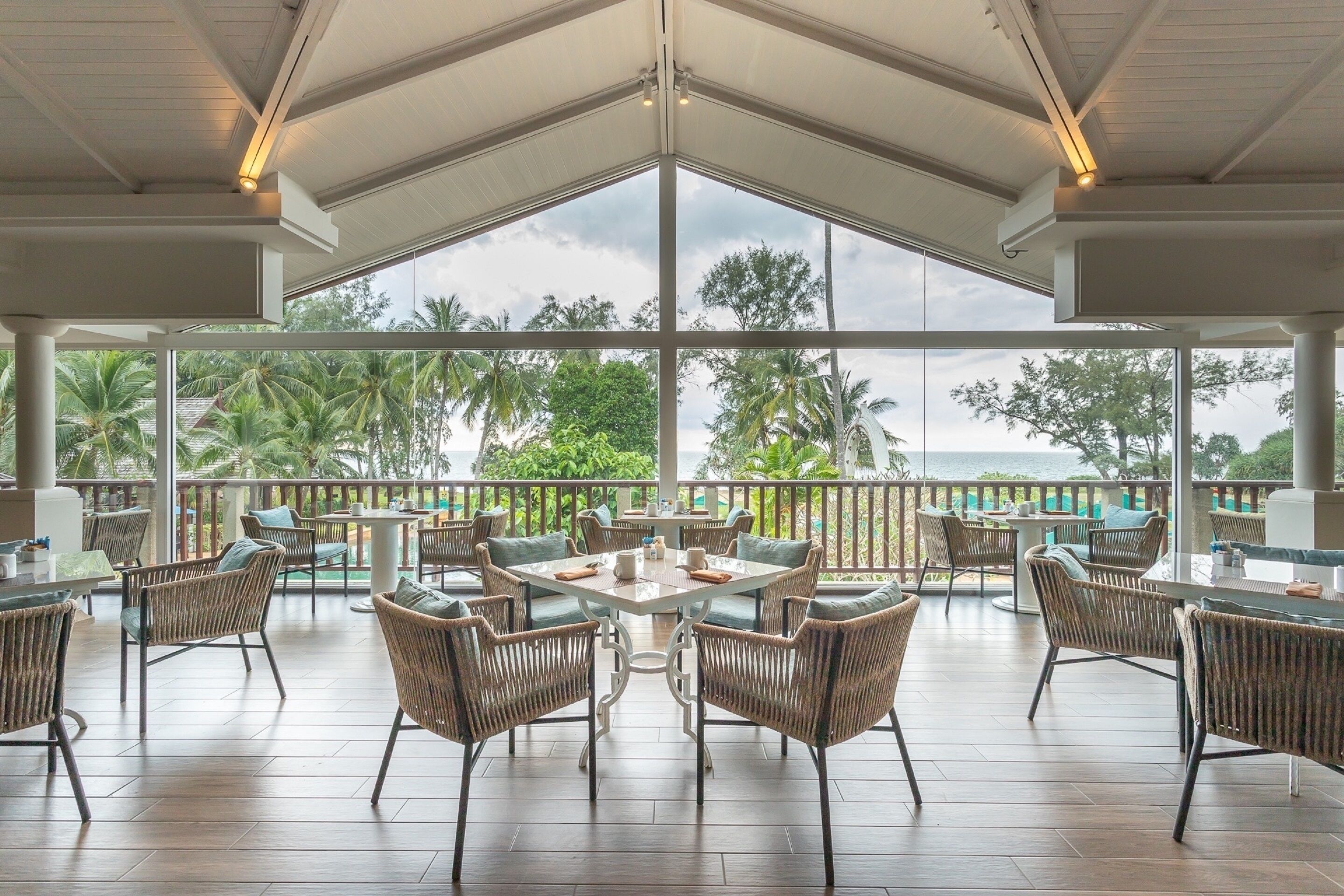 Un amplio espacio al aire libre con mobiliario de mimbre y una mesa grande, con vistas panorámicas a palmeras y al mar.