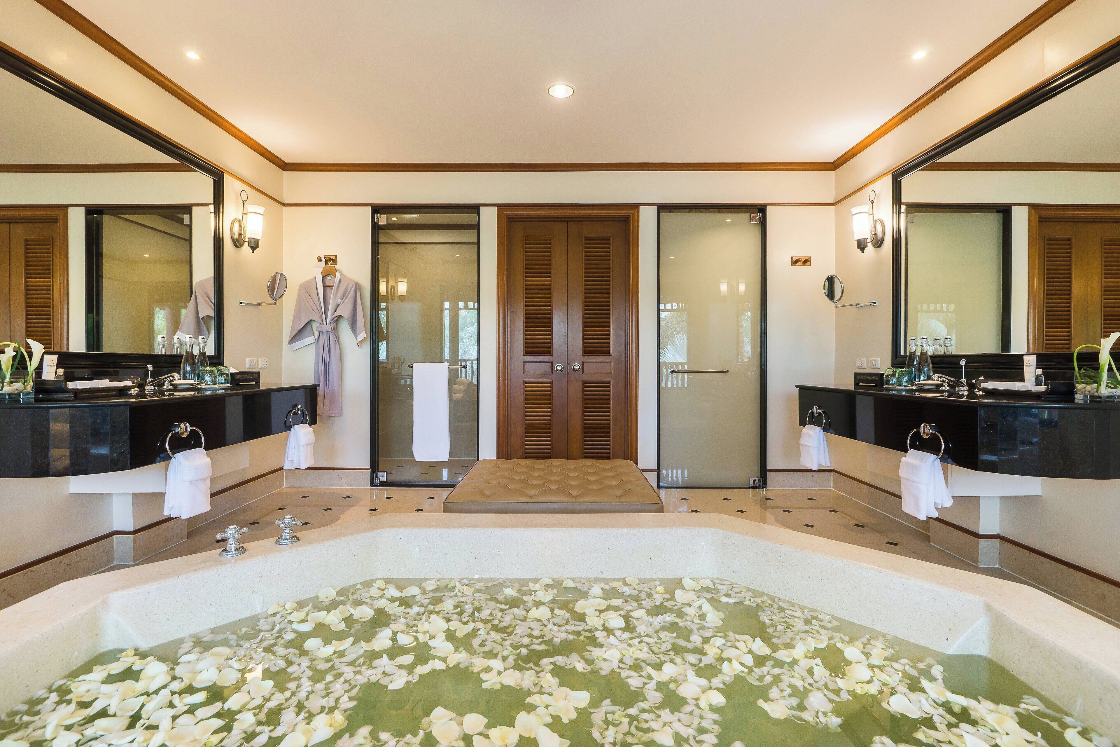 A spacious bathroom with a large bathtub filled with rose petals, two sinks with modern fixtures, and a large mirror above each sink.