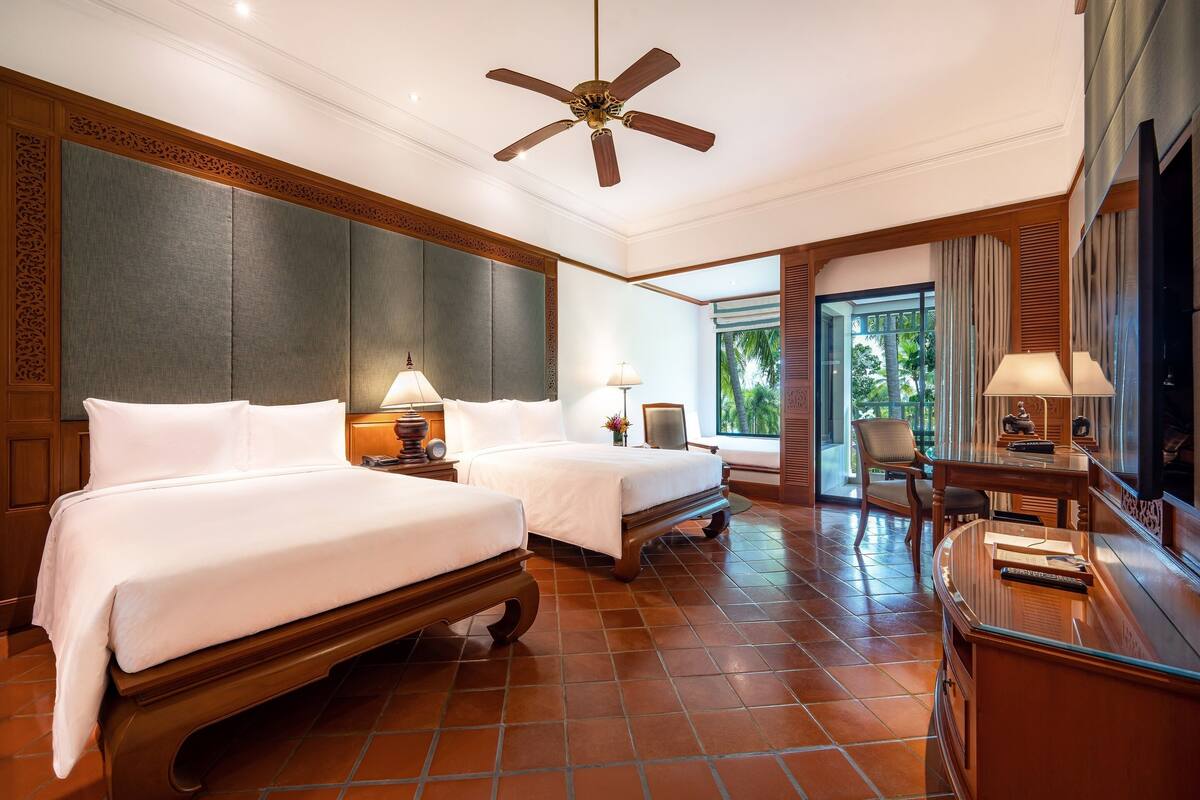 A hotel room with two beds, a desk, a chair, a ceiling fan, and a large window with a view of palm trees.