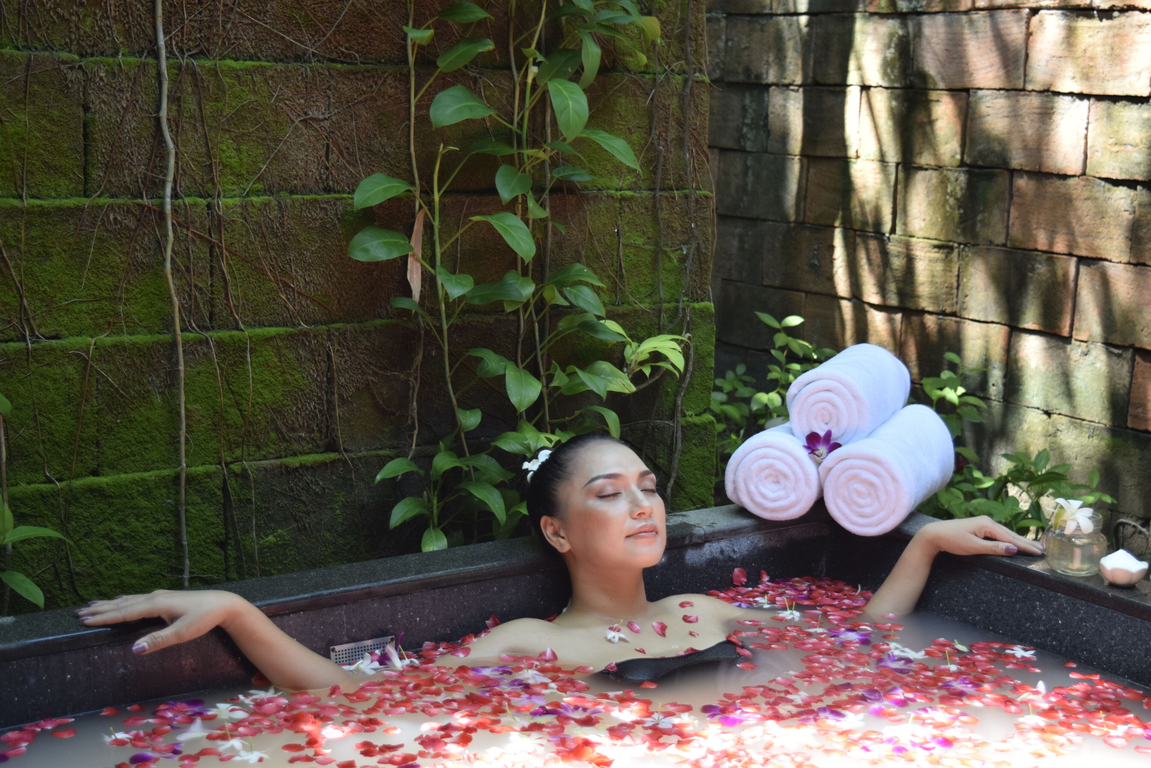 A person relaxing in a bathtub filled with rose petals, surrounded by greenery and a brick wall.