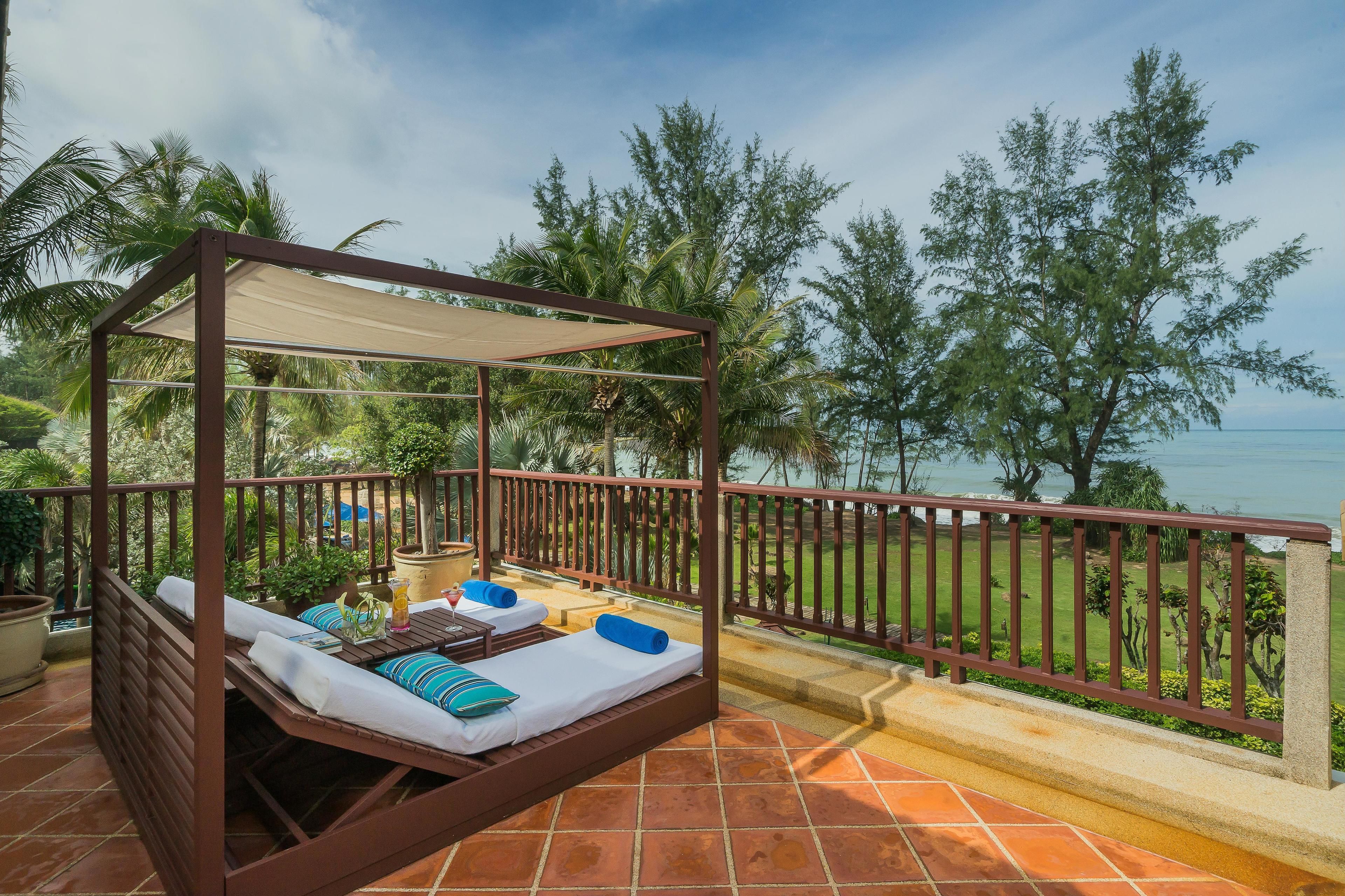 A wooden four-poster bed with white linens and blue pillows, a small table with a drink, and a view of the sea and palm trees.