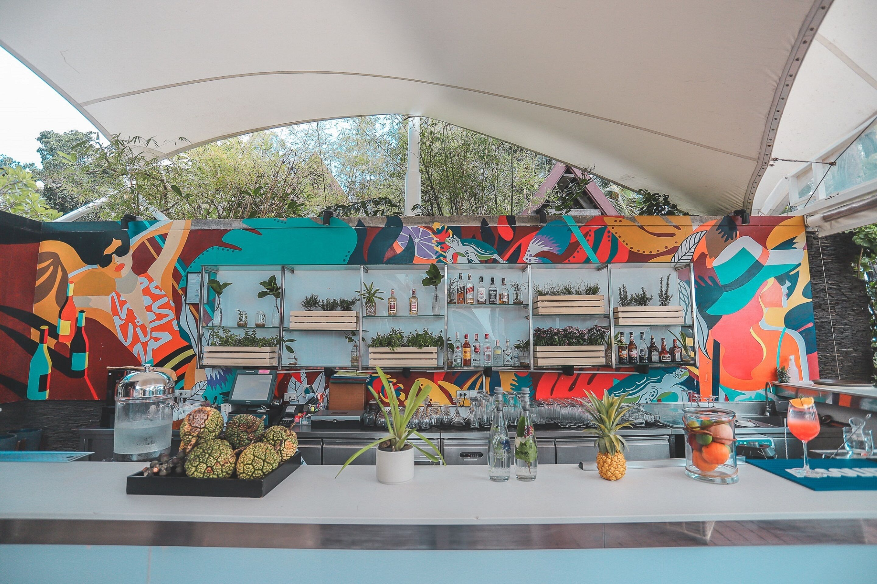 Zona de bar con murales coloridos, estantes llenos de botellas y una exhibición de frutas.