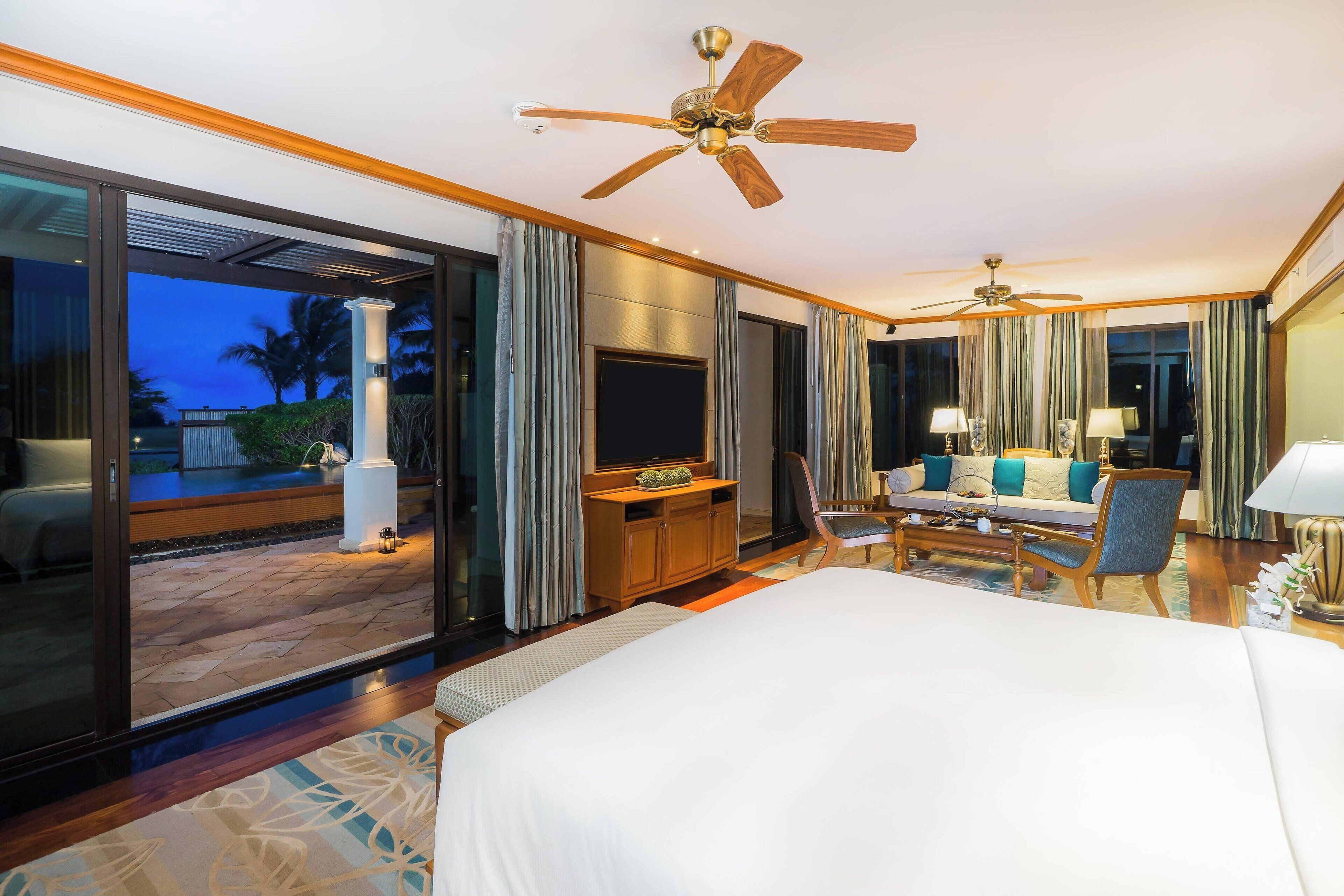 Una habitación de hotel con una cama grande, televisión, sofá, sillón y ventilador de techo.
