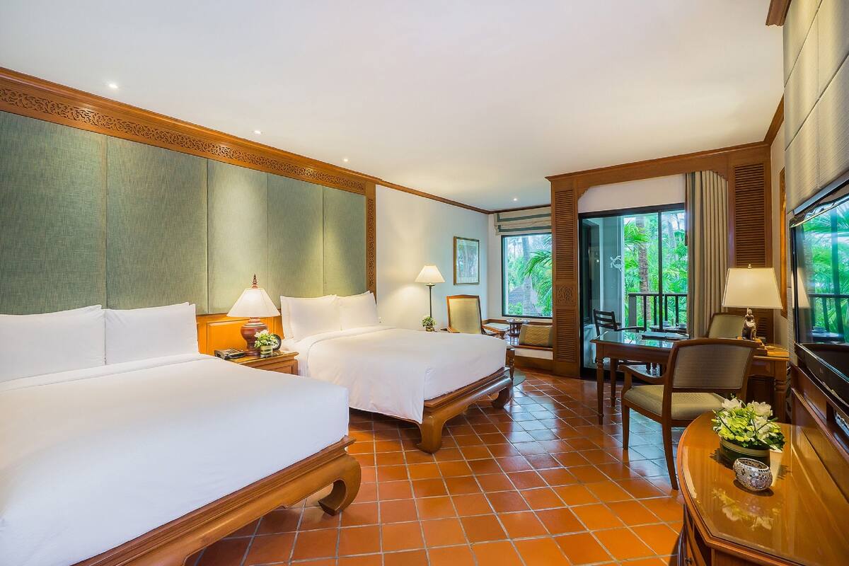 A hotel room with two beds, a dining table, chairs, a TV, and a window with a view of greenery.