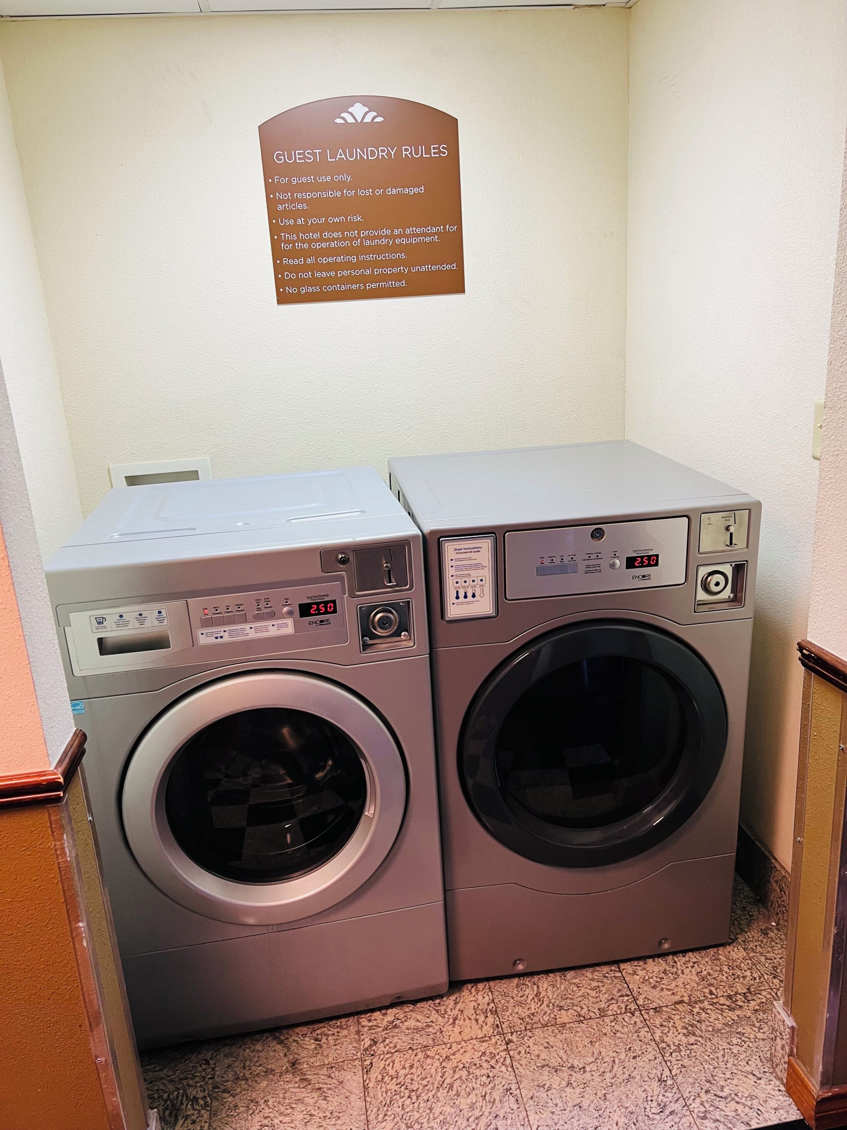 A pair of front-loading washing machines in a hotel room, with a sign above them detailing guest laundry rules.