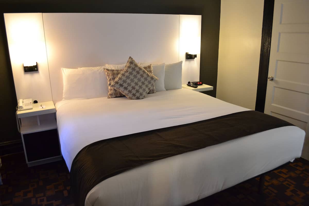 Una cama bien hecha con sábanas blancas y una manta oscura, una mesita de noche con lámpara, y un reloj de pared.