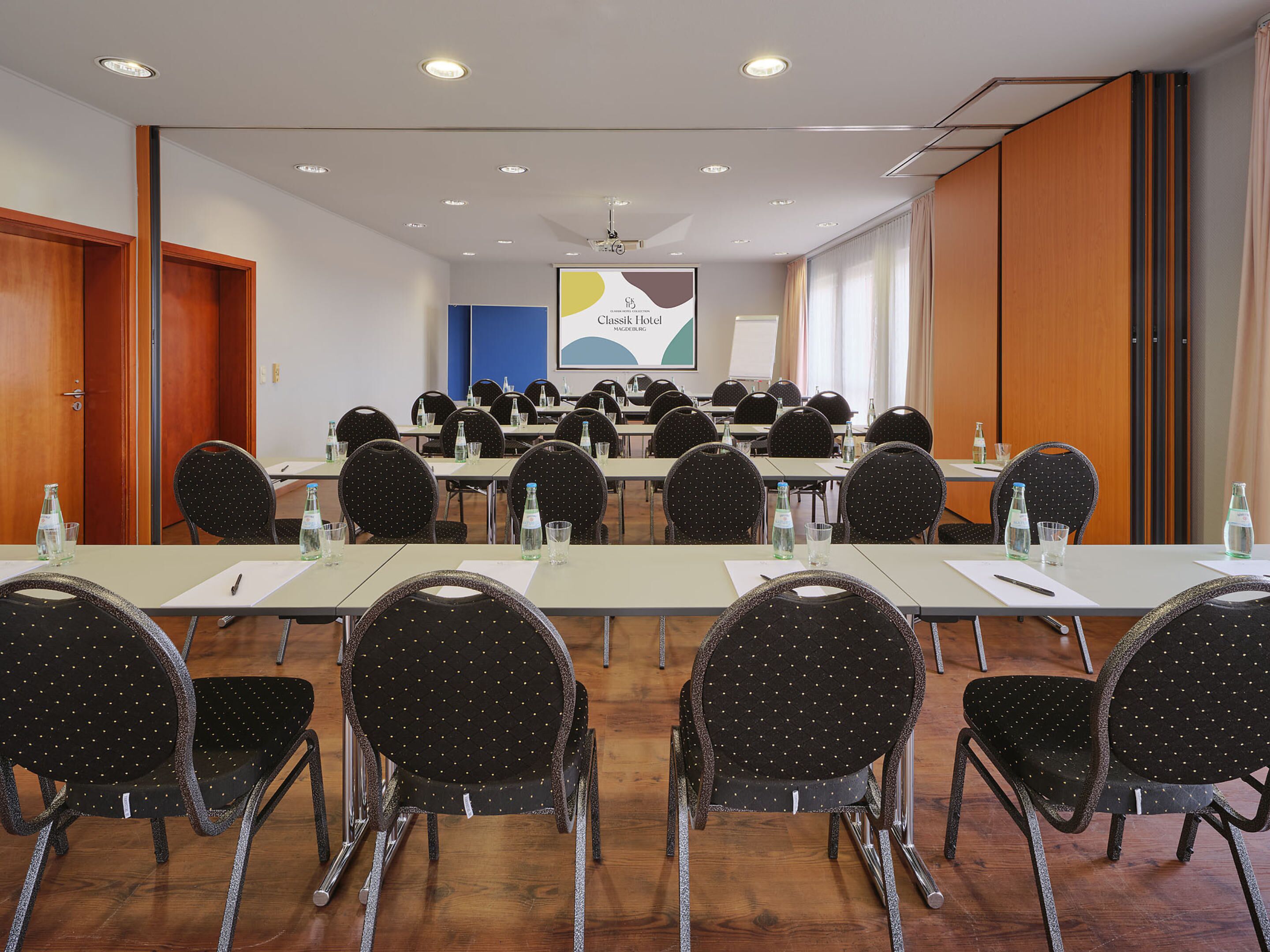 A conference room with multiple tables and chairs, equipped with water bottles and papers, in a hotel setting.