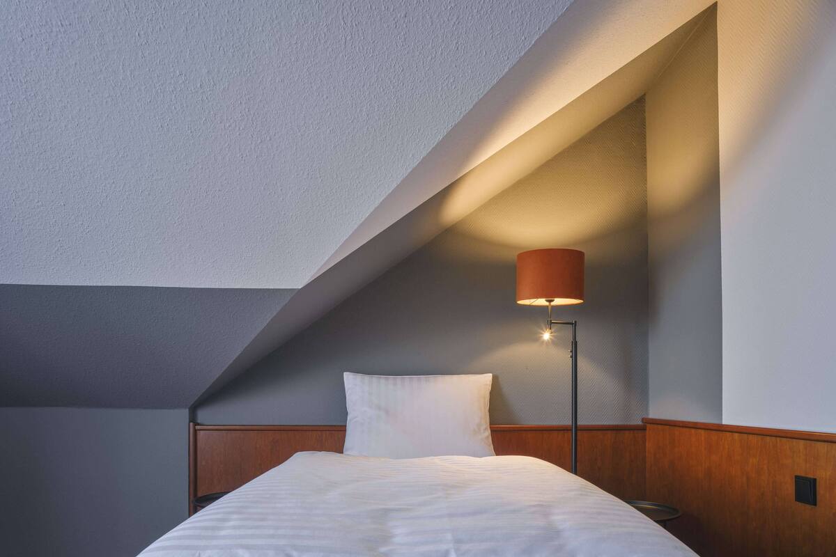 Habitación de hotel con techo inclinado, una cama individual, una mesita de noche y una lámpara de pie.