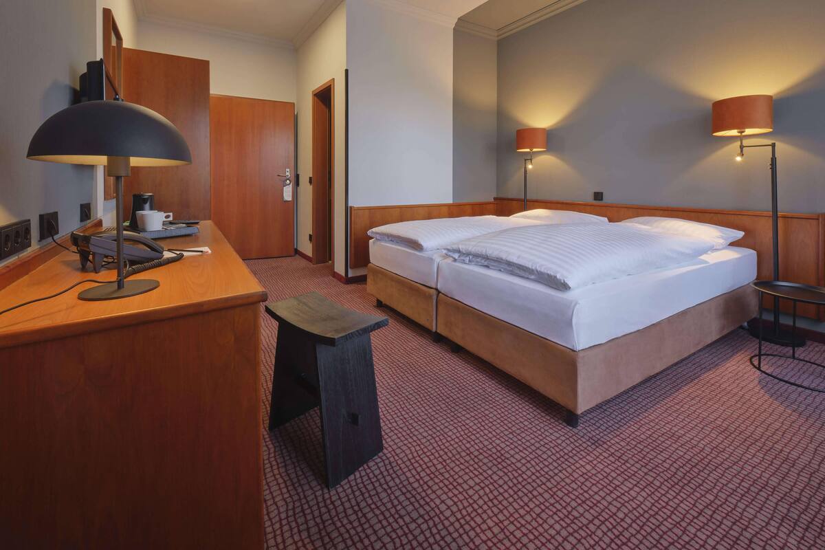 Habitación de hotel con escritorio de madera, teléfono, taburete pequeño, cama doble con sábanas blancas, dos lámparas fijadas a la pared y puerta que da a otra habitación.