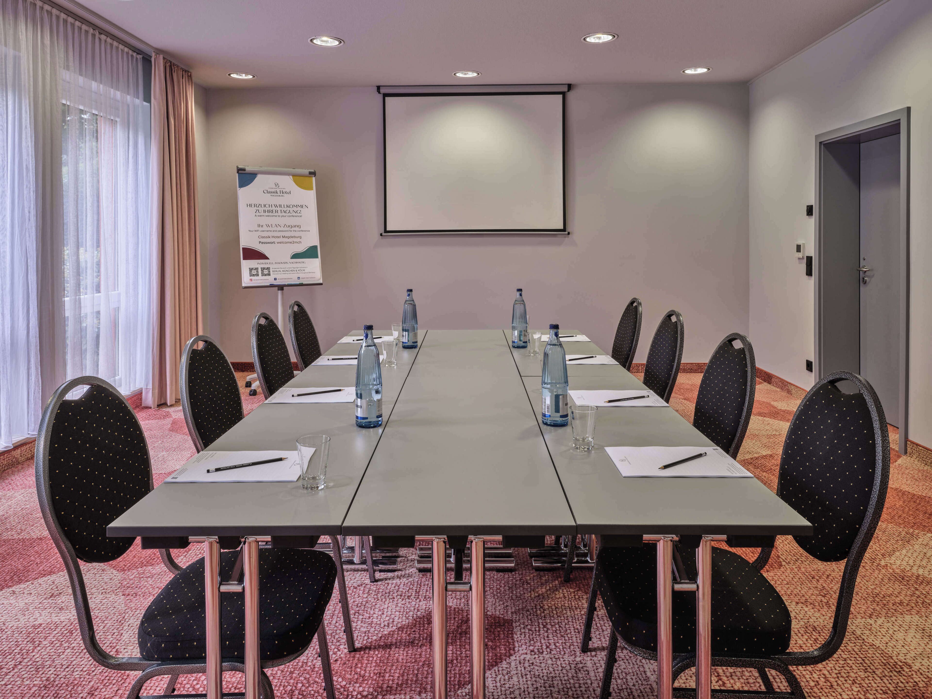 Un salón de conferencias con una larga mesa, sillas, botellas de agua y una pantalla de proyector.