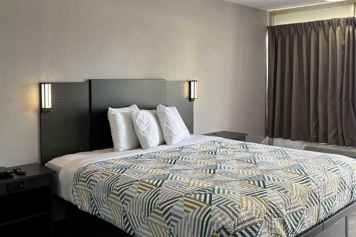 A hotel room with a bed, two bedside tables, a wall-mounted light, a window with curtains, and a built-in air conditioning unit.