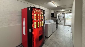 A vending machine with various drink options, a sink, and a refrigerator in a hallway with white brick walls and tiled flooring. - Motel 6 Richmond, KY – EKU Campus Area (Richmond)
