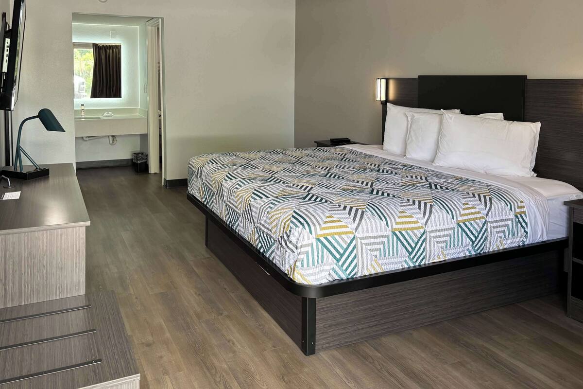 A modern hotel room with a large bed, a desk with a lamp, and a bathroom visible through an open door.