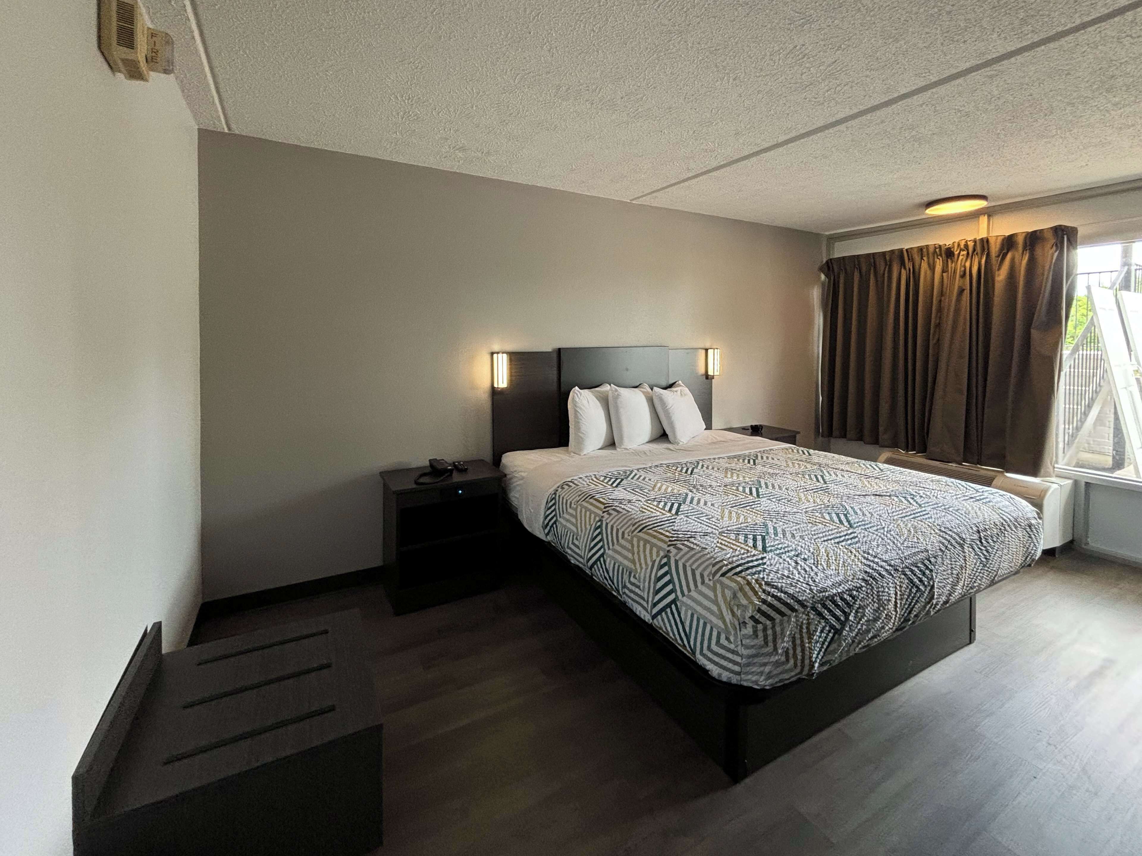A modern hotel room with a large bed, a nightstand, a desk, and a window with curtains.