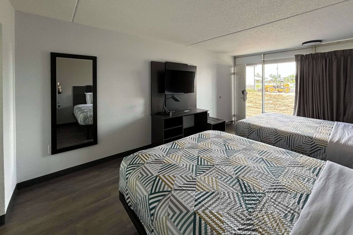 A hotel room with two beds, a TV, a mirror, and a window with curtains.