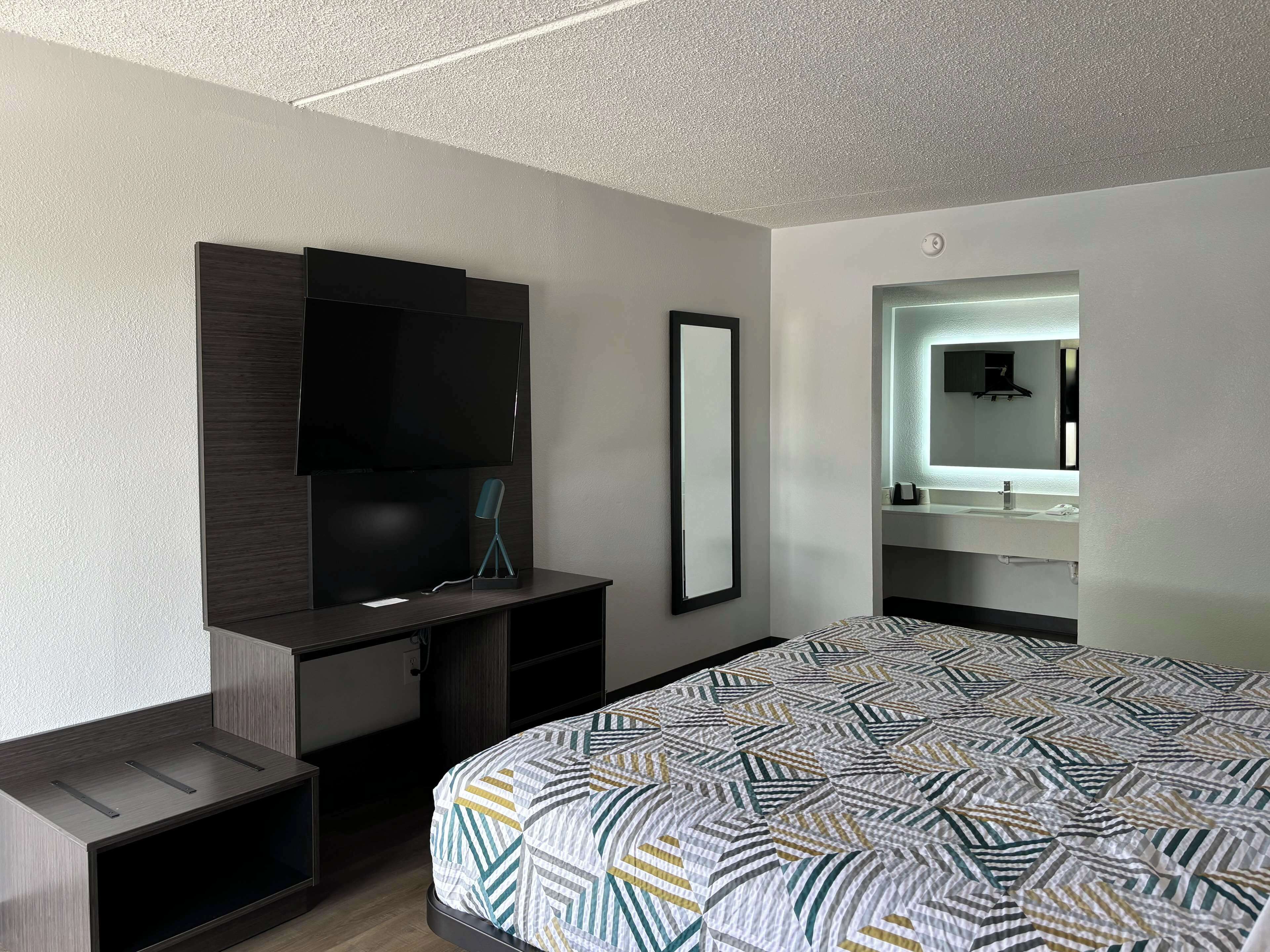 Quarto de hotel moderno com uma cama grande, uma mesa de trabalho com televisão de tela plana, um espelho e uma toucador embutida com pia.