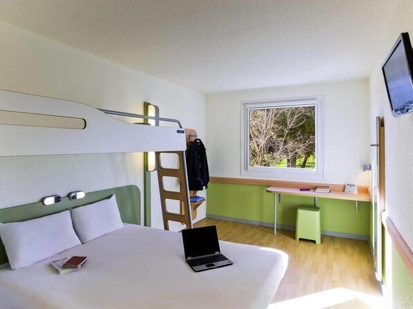 Une chambre d’hôtel avec un lit superposé, un bureau avec une chaise, un ordinateur portable, une télévision et une fenêtre donnant sur des arbres.
