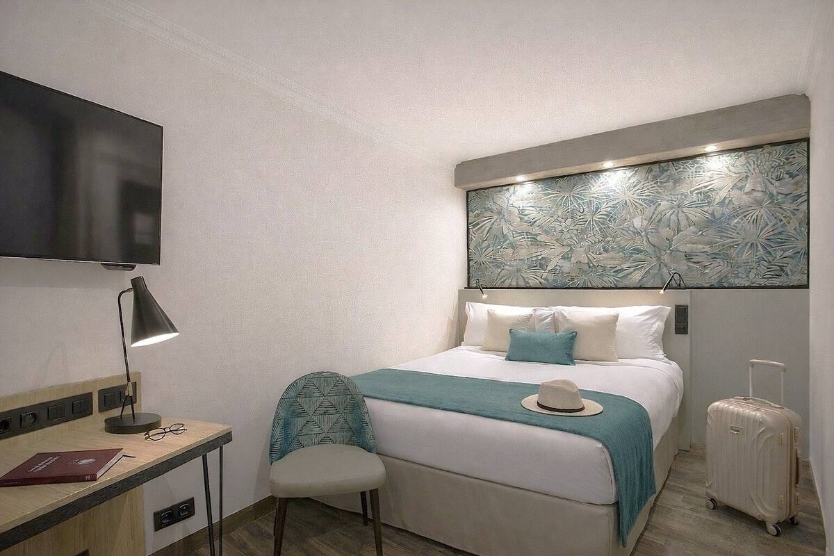 Habitación de hotel compacta con cama, escritorio con lámpara, silla, maleta y televisor montado en la pared.