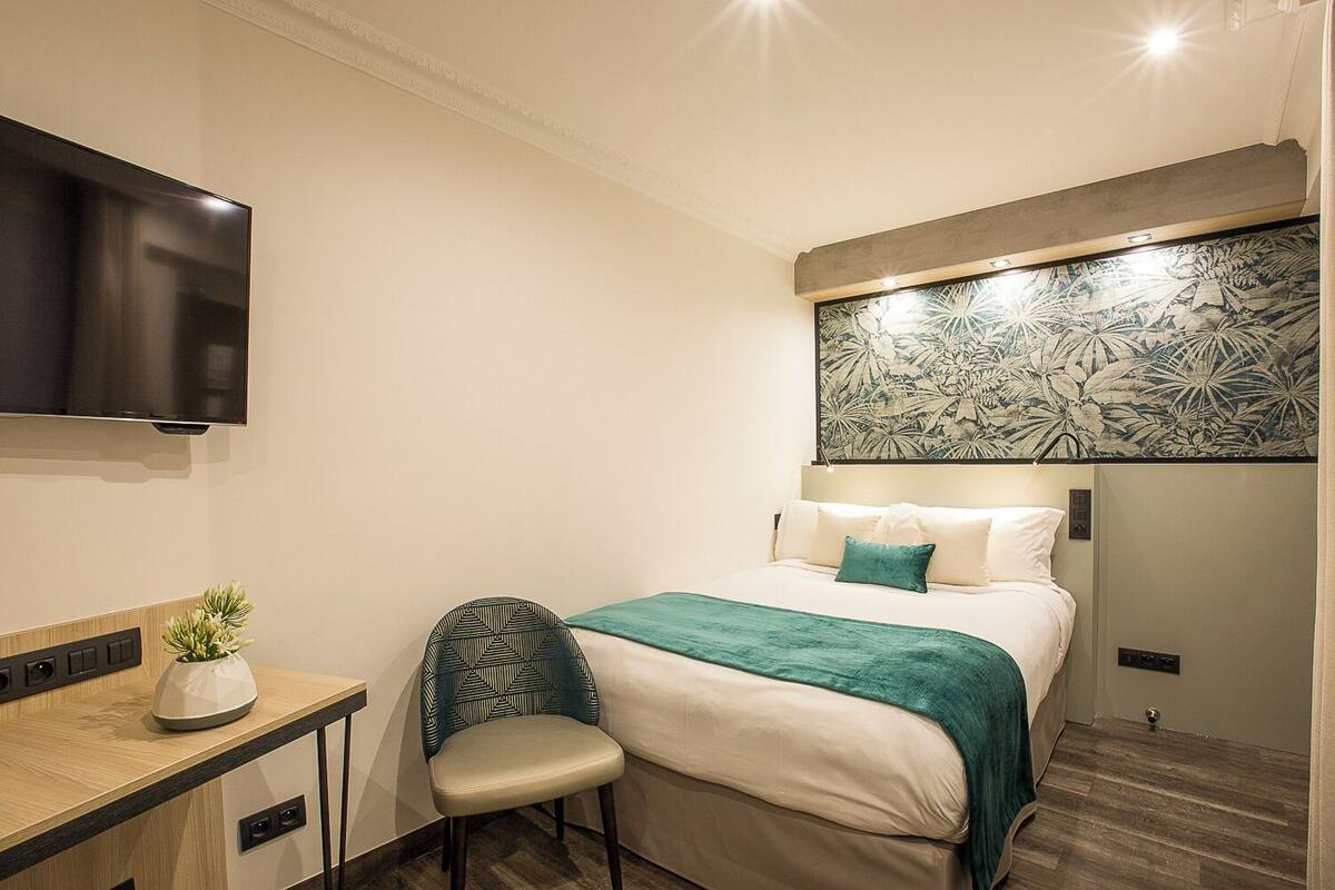 Habitación de hotel con cama, escritorio con planta, silla, televisor y un panel decorativo en la pared.