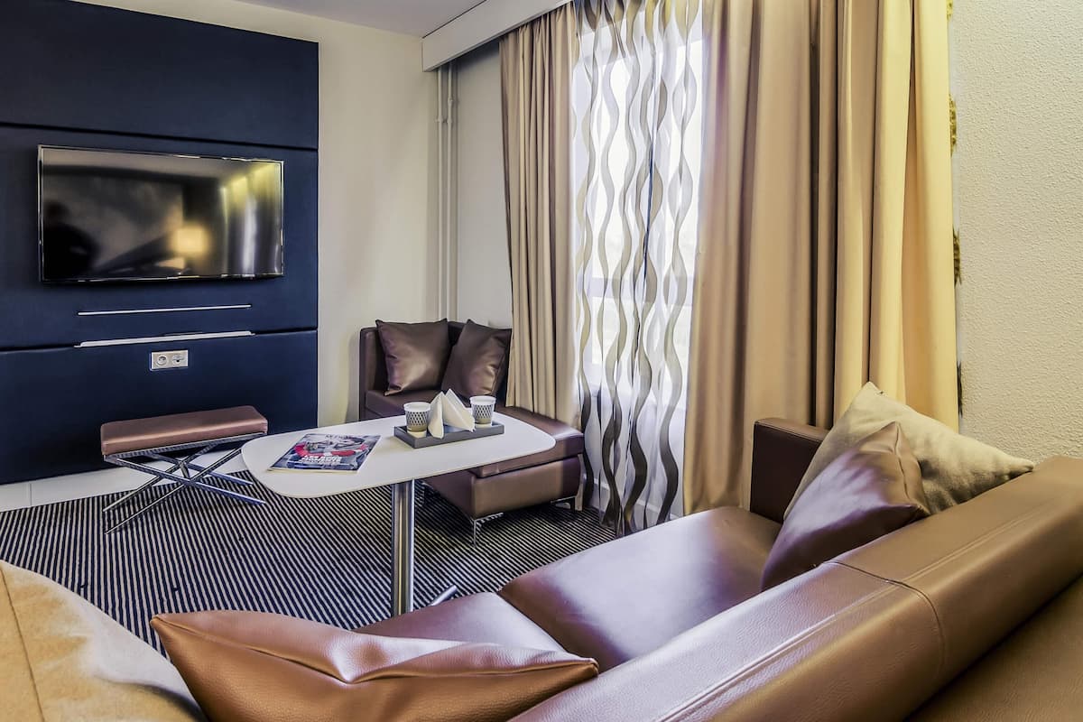 A modern hotel room with a flat-screen TV mounted on the wall, a sofa, a coffee table, and a patterned curtain.