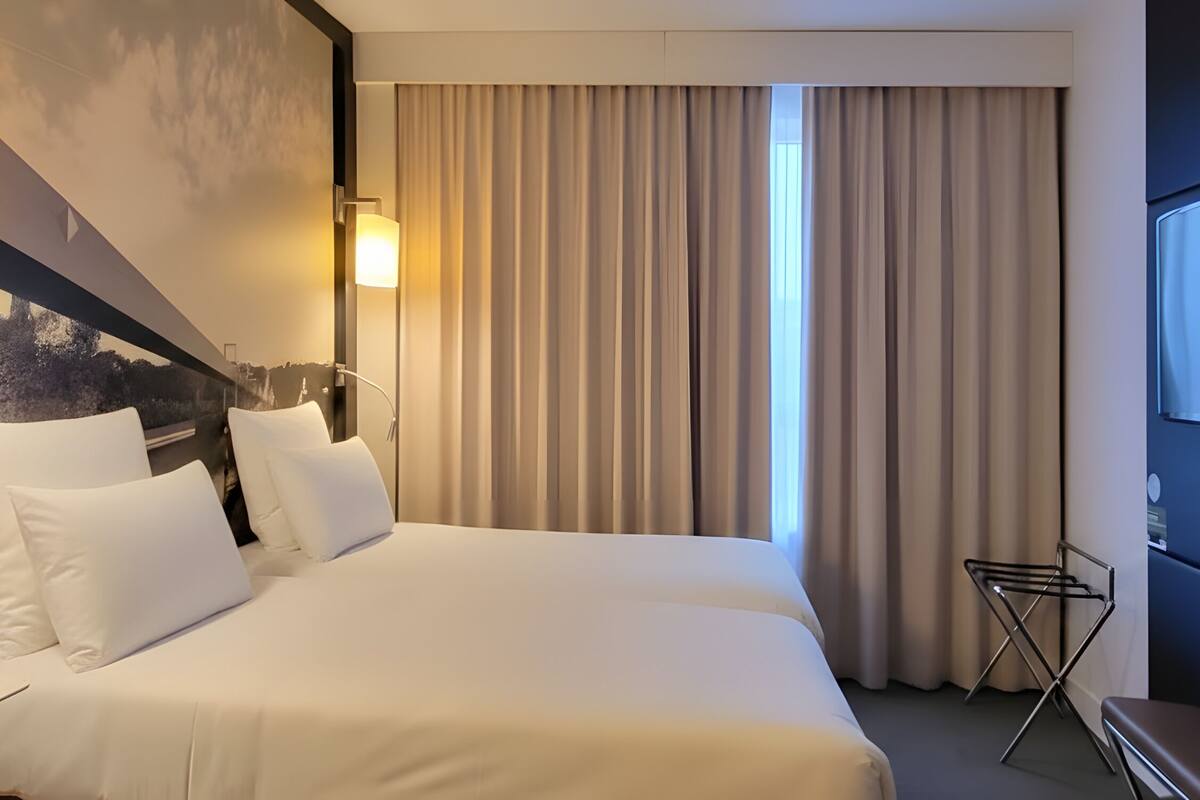 A hotel room with a bed, bedside table with a lamp, a TV mounted on the wall, and a window with curtains.