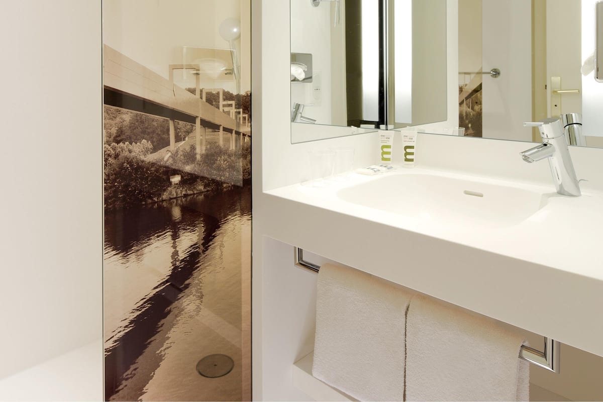 A modern bathroom with a white sink, chrome faucet, and a large mirror. There is a towel rack and a glass shower door.
