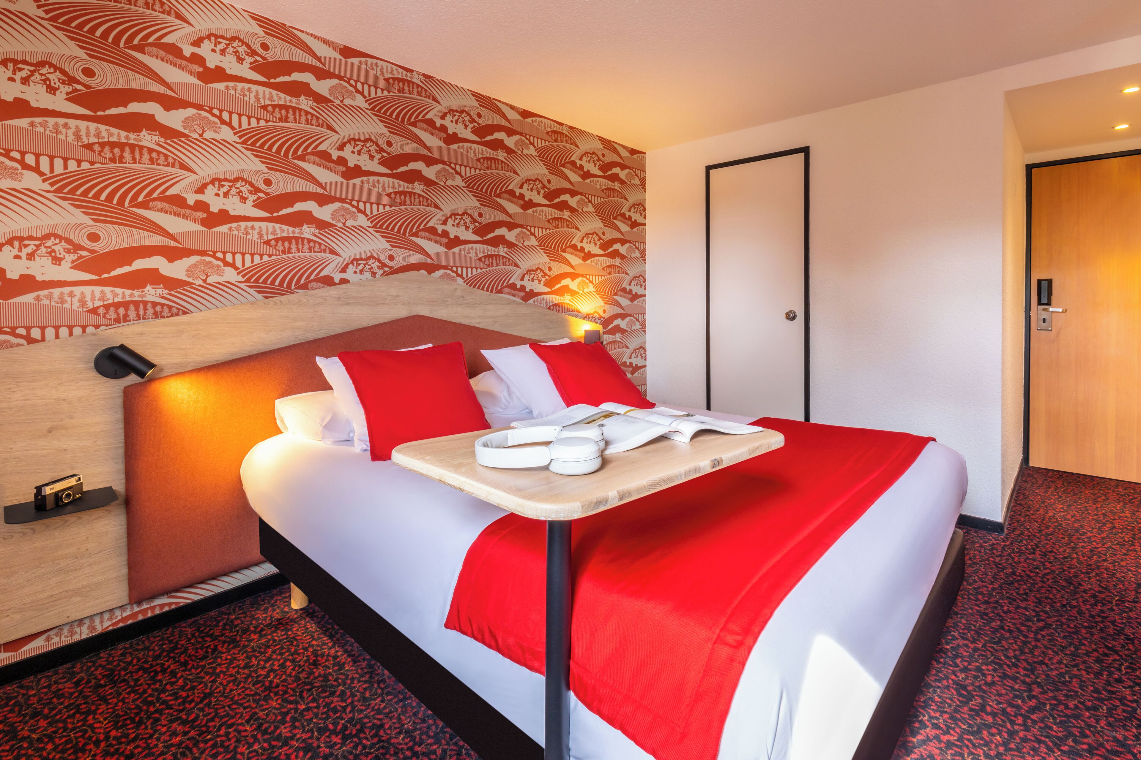 A hotel room with a bed, a wooden headboard, a red and white bedspread, a bedside table with a lamp, and a red carpeted floor.