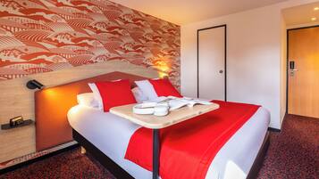 A hotel room with a bed, a wooden headboard, a red and white bedspread, a bedside table with a lamp, and a red carpeted floor.