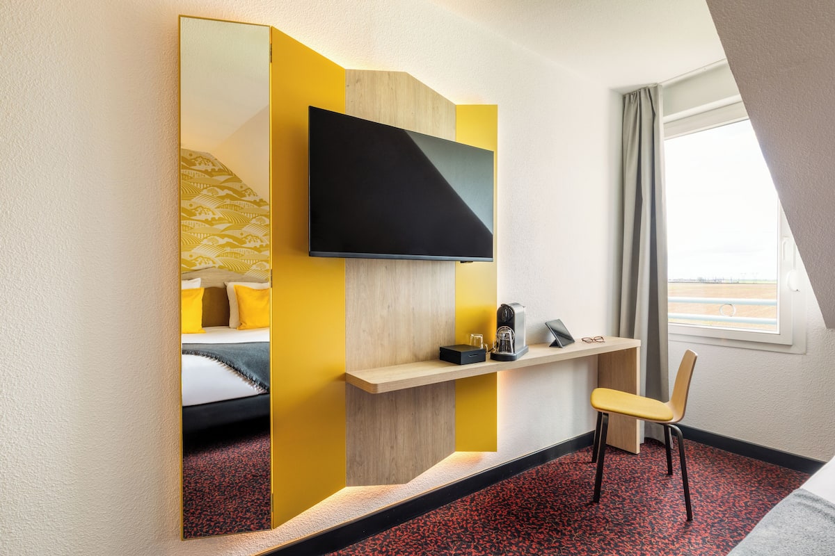 Une chambre d’hôtel avec un lit, un téléviseur fixé au mur, un bureau avec une chaise et une fenêtre avec des rideaux.