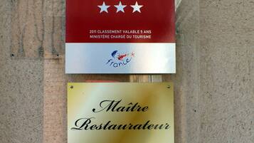 Un letrero de hotel con un logotipo rojo y blanco, una placa dorada con la leyenda 'Maitre Restaurateur' y una placa de matrícula que indica 'IV LICENCE du 24 Septembre 1941'.