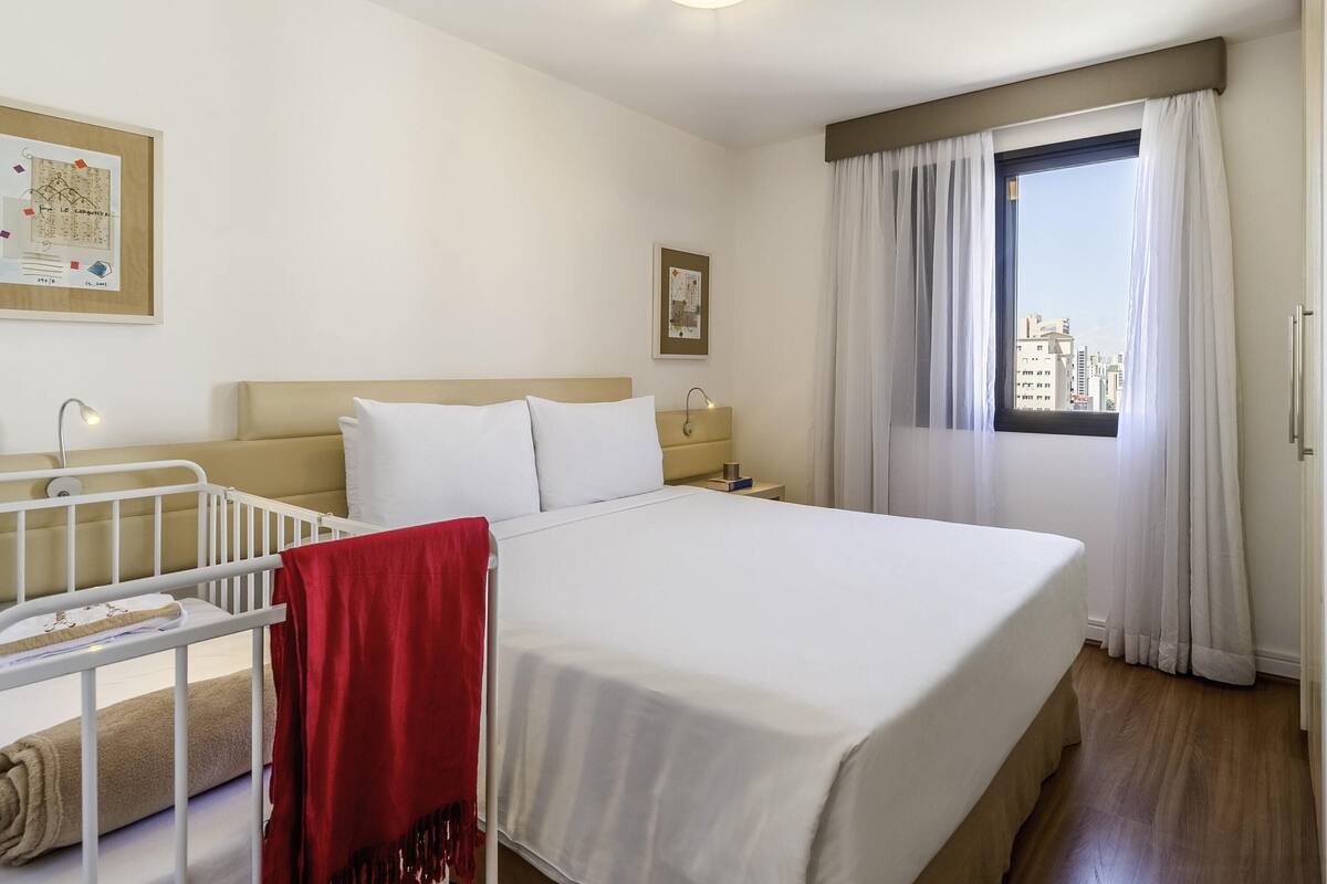 Ein Hotelzimmer mit einem Bett, einem Babybett, einem Nachttisch und einem Fenster mit Vorhängen.