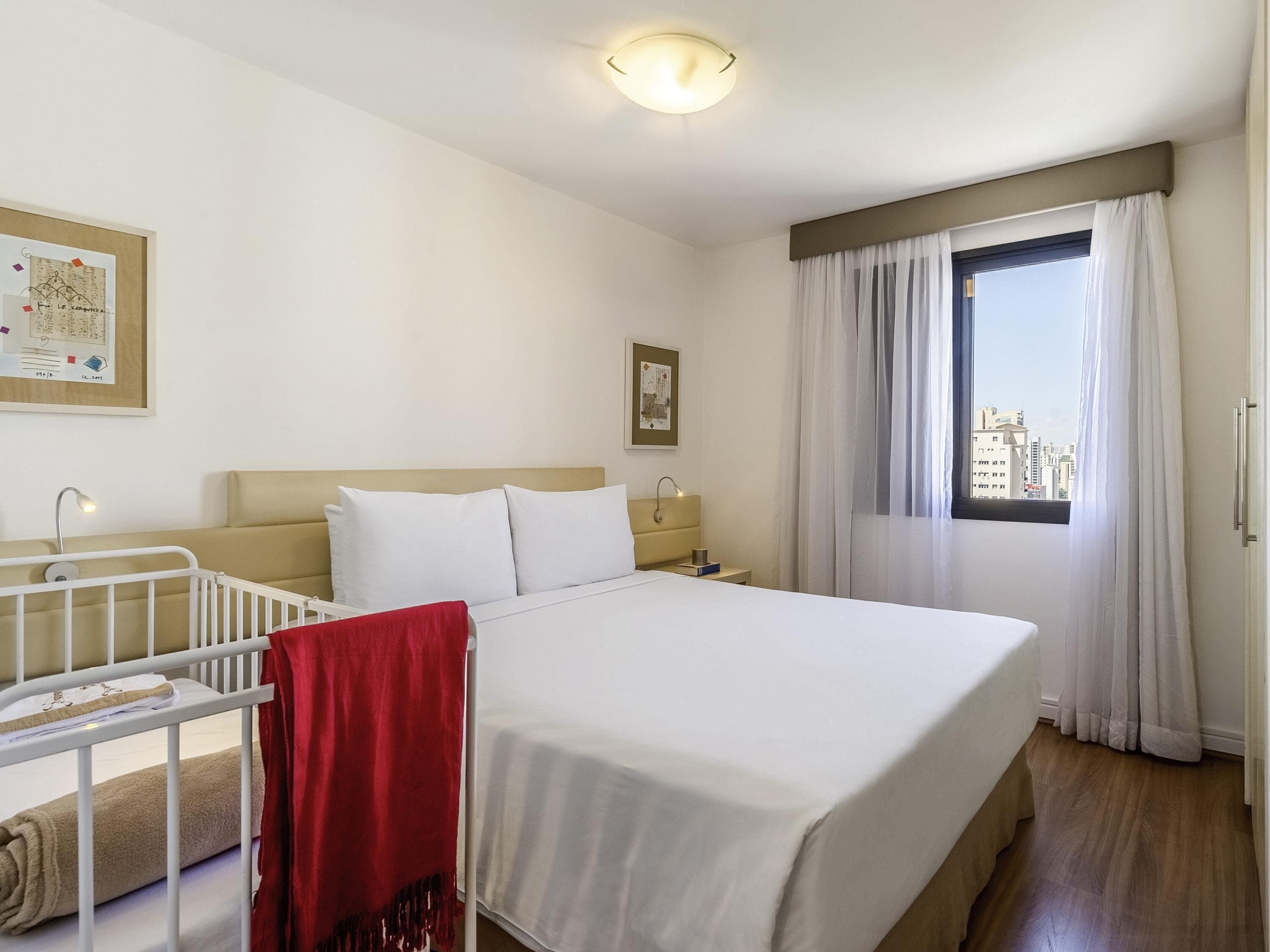 Una camera d'hotel con un letto, una culla, un comodino, una lampada e una finestra con tende.