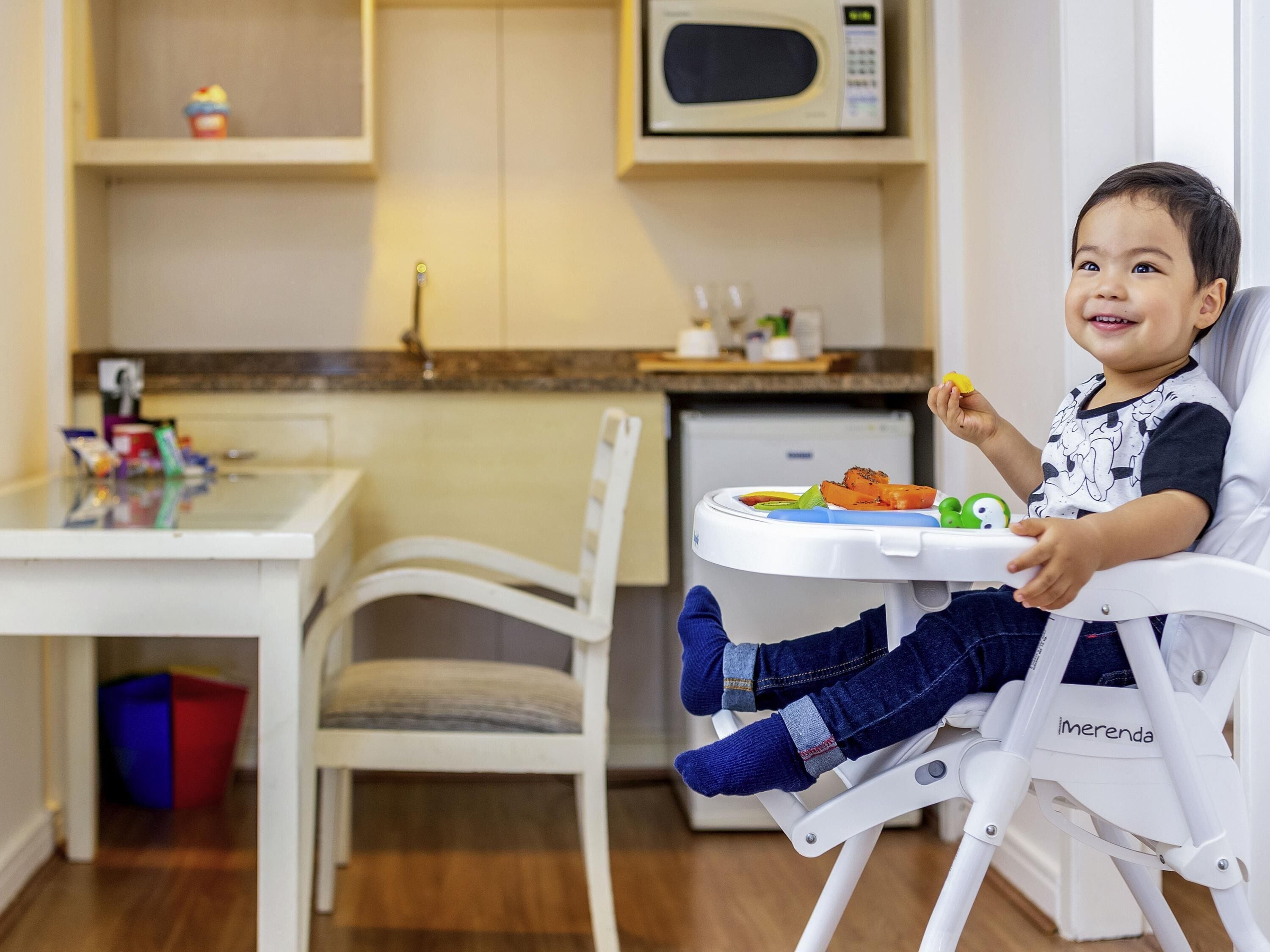 Un bambino seduto in una seggiolina da pranzo con un vassoio, un forno a microonde sopra e un piano di lavoro con diversi oggetti.