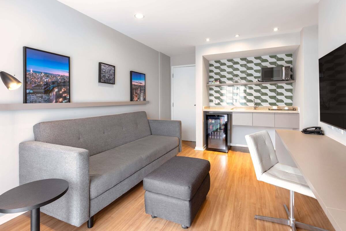 Ein modernes Wohnzimmer mit einem grauen Sofa, einem kleinen runden Tisch und einem weißen Stuhl. Es gibt eine Küchenzeile mit einer Mikrowelle und einer gemusterten Spritzschutzwand.