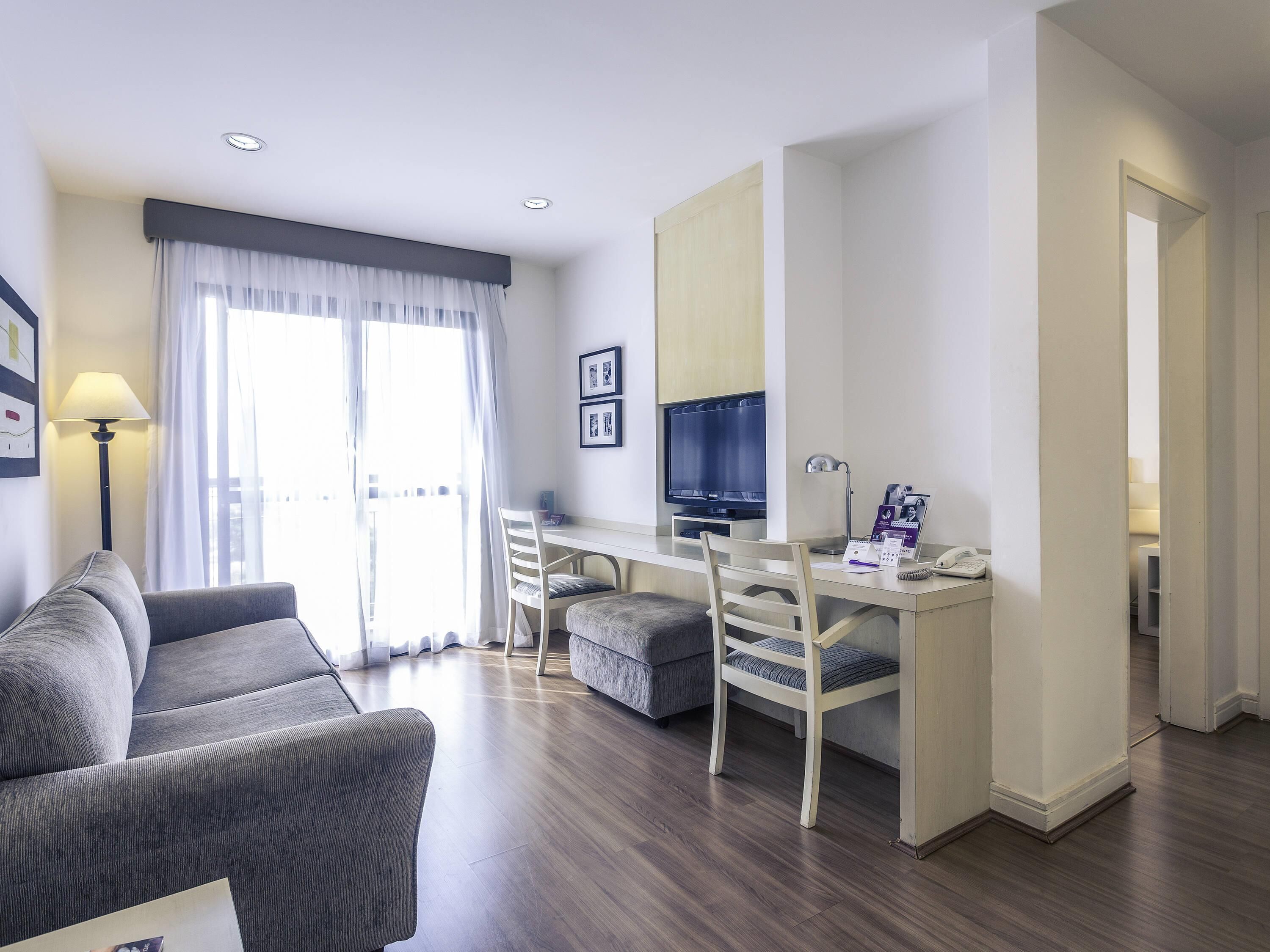 Une chambre d’hôtel moderne équipée d’un canapé, d’un bureau avec un ordinateur, d’une télévision et d’une fenêtre avec des rideaux.