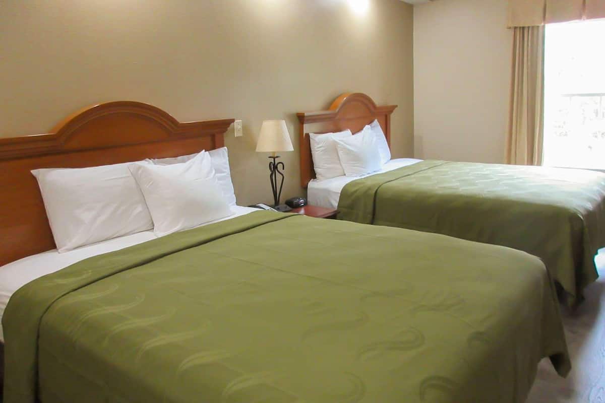 A hotel room with two beds, each with a wooden headboard, a bedside table with a lamp, and a window with curtains.