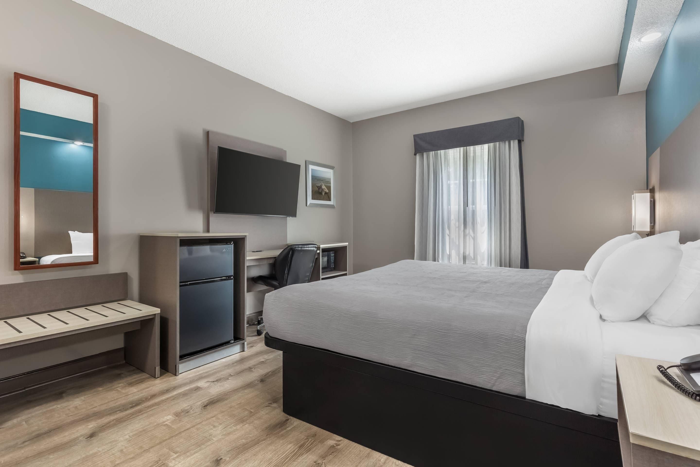 A hotel room with a bed, a desk with a telephone, a television, a mirror, a small refrigerator, and a window with curtains.