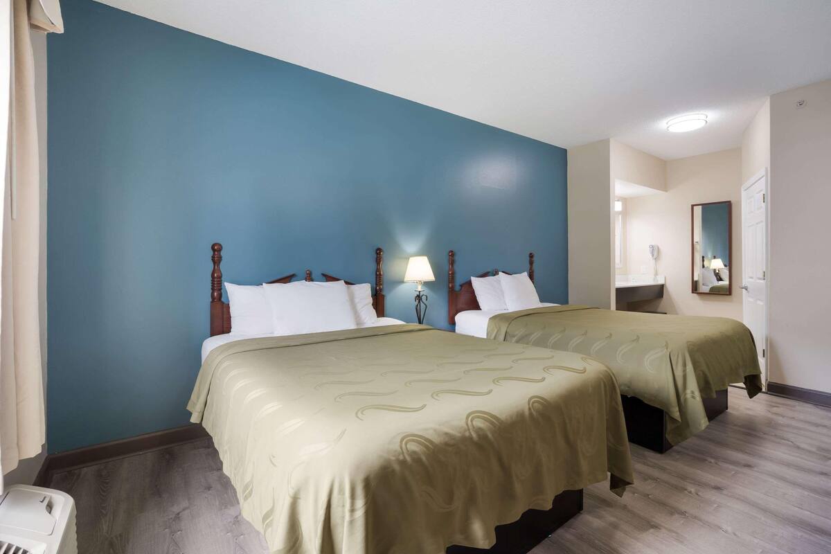 A hotel room with two beds, a blue accent wall, a wooden headboard, a bedside lamp, and a mirror.