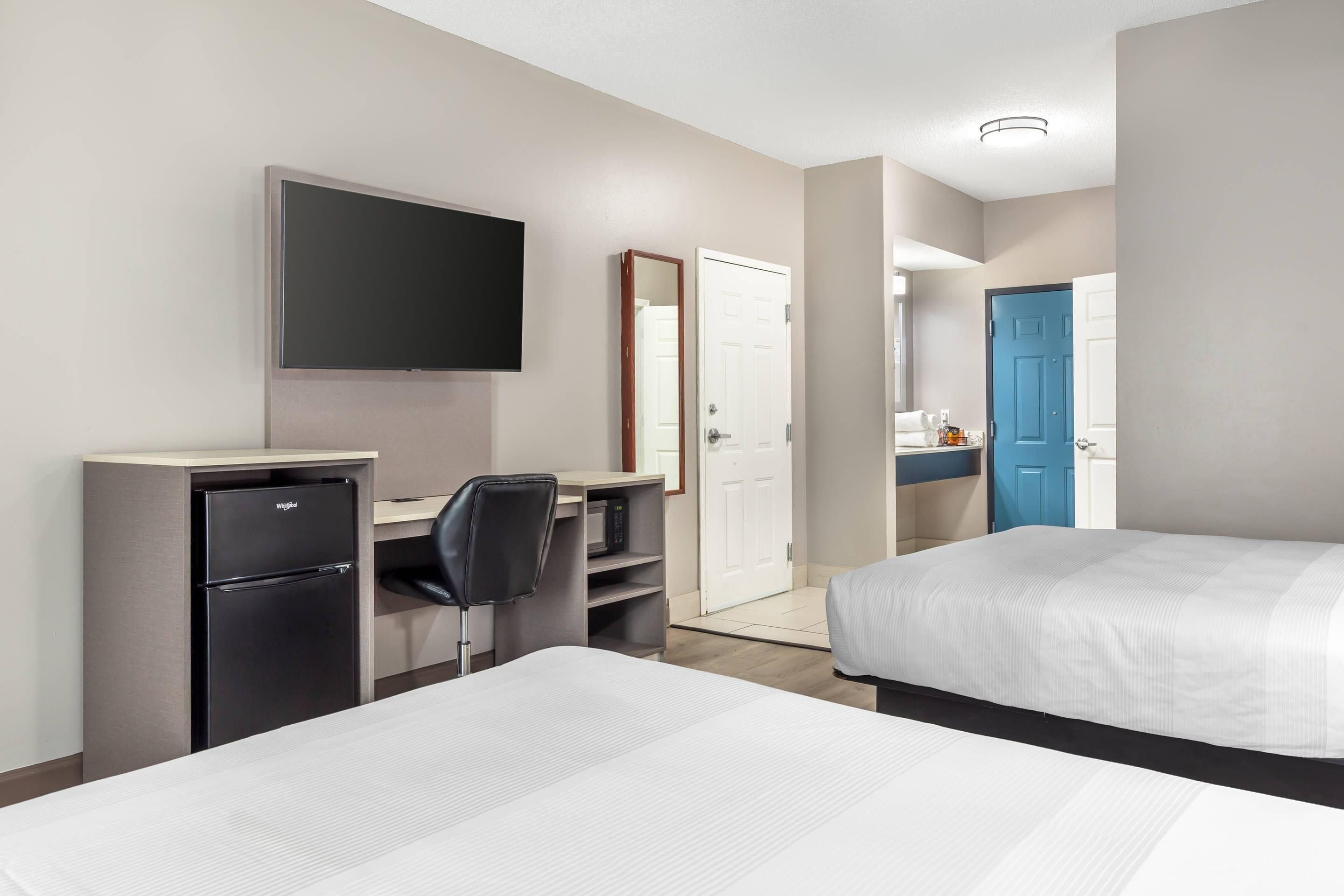 A hotel room with a bed, a desk with a computer, a refrigerator, a television, and a bathroom with a blue door.