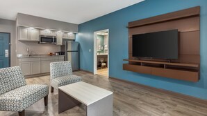 A modern living room with a flat-screen TV, two patterned armchairs, a coffee table, and a kitchen area with built-in appliances.