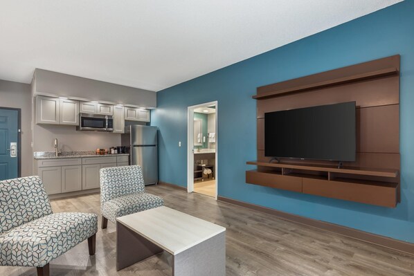 A modern living room with a flat-screen TV, two patterned armchairs, a coffee table, and a kitchen area with built-in appliances.
