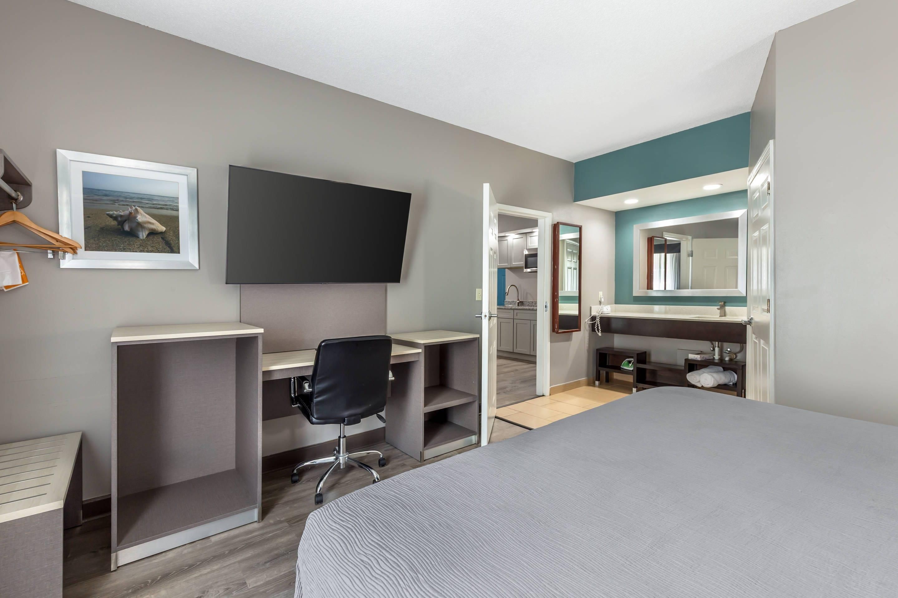 A hotel room with a bed, desk, chair, television, and a bathroom with a mirror and sink.