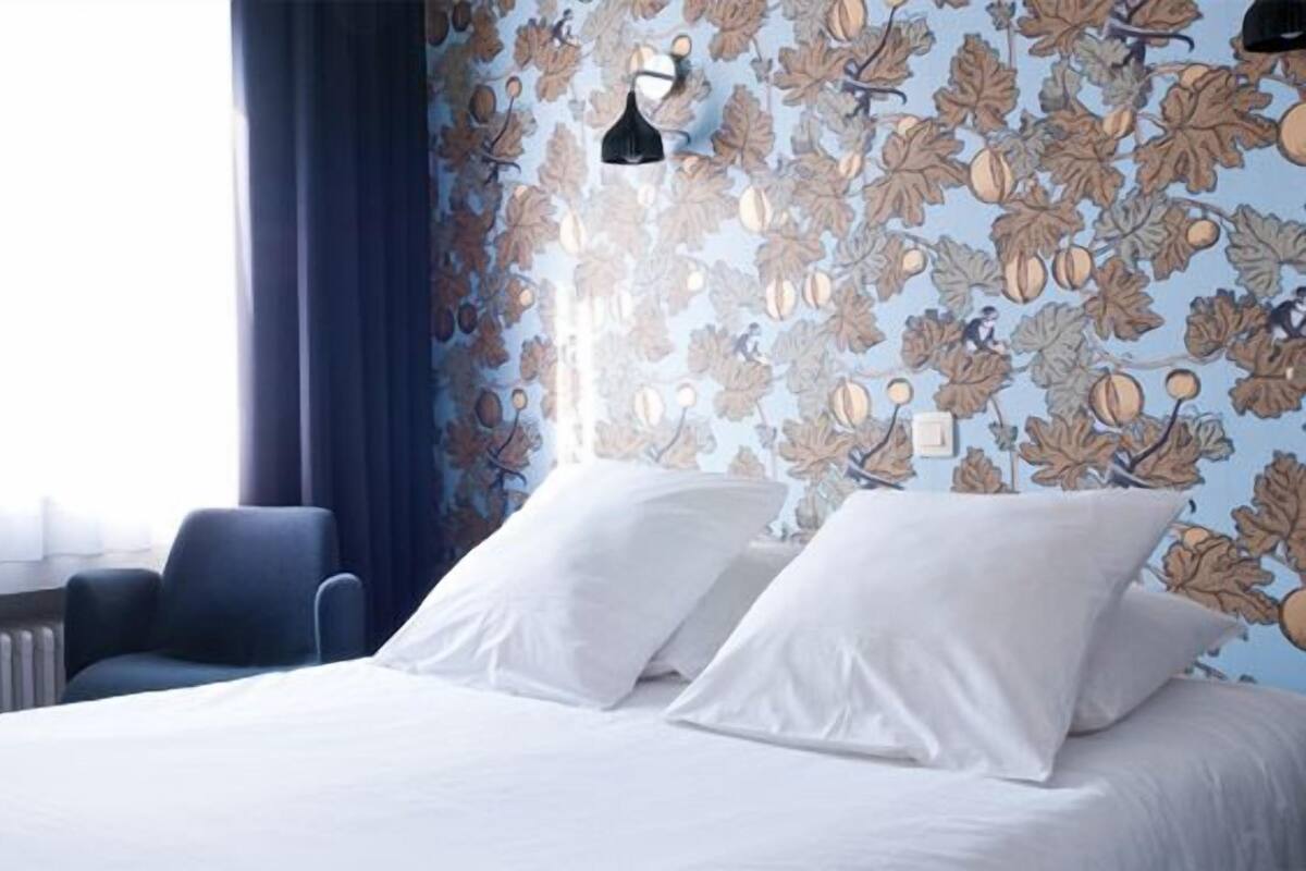 A hotel room with a bed, white pillows, a blue chair, and a wall with a leaf pattern.