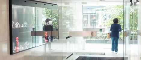 A modern lobby with a glass entrance, a reception desk, and a visible outdoor area with greenery.